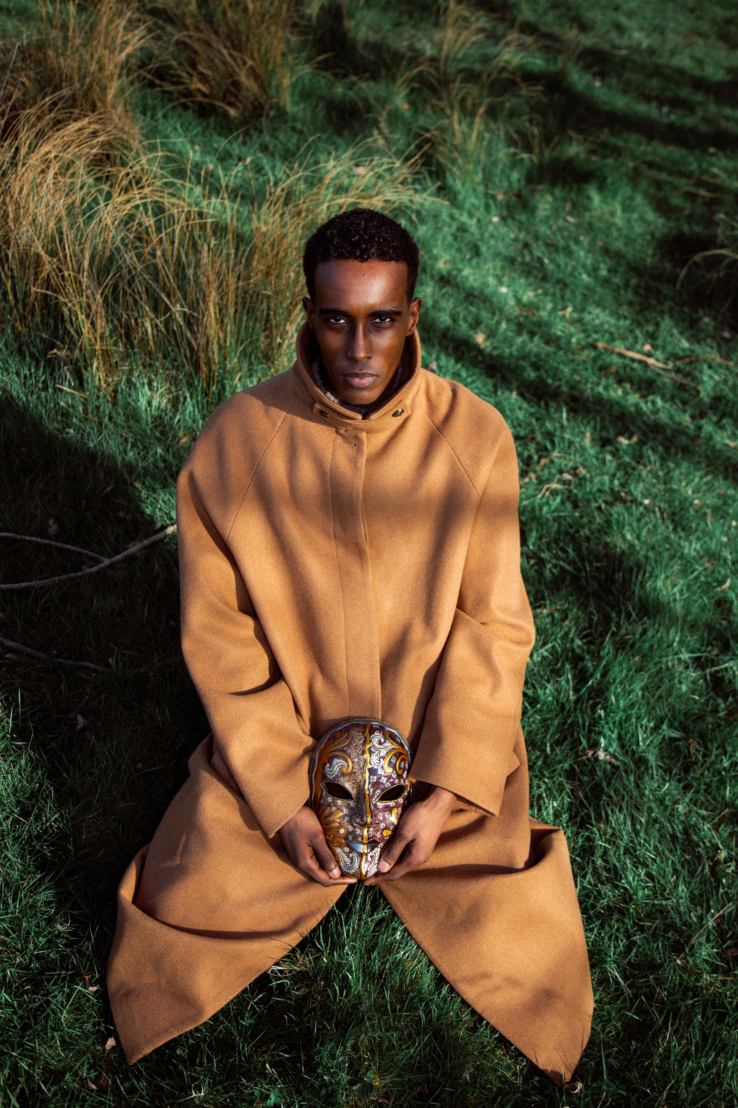 Man in tan coat kneeling on grass, holding ornate golden skull mask in outdoor setting.