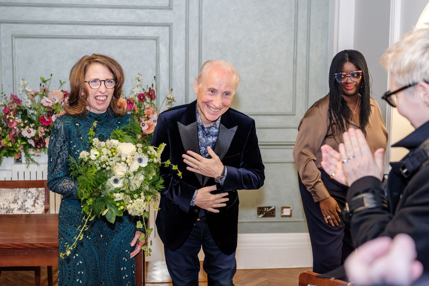 Two grooms exchange vows at a same-sex wedding ceremony on a checkered floor with guests applauding.