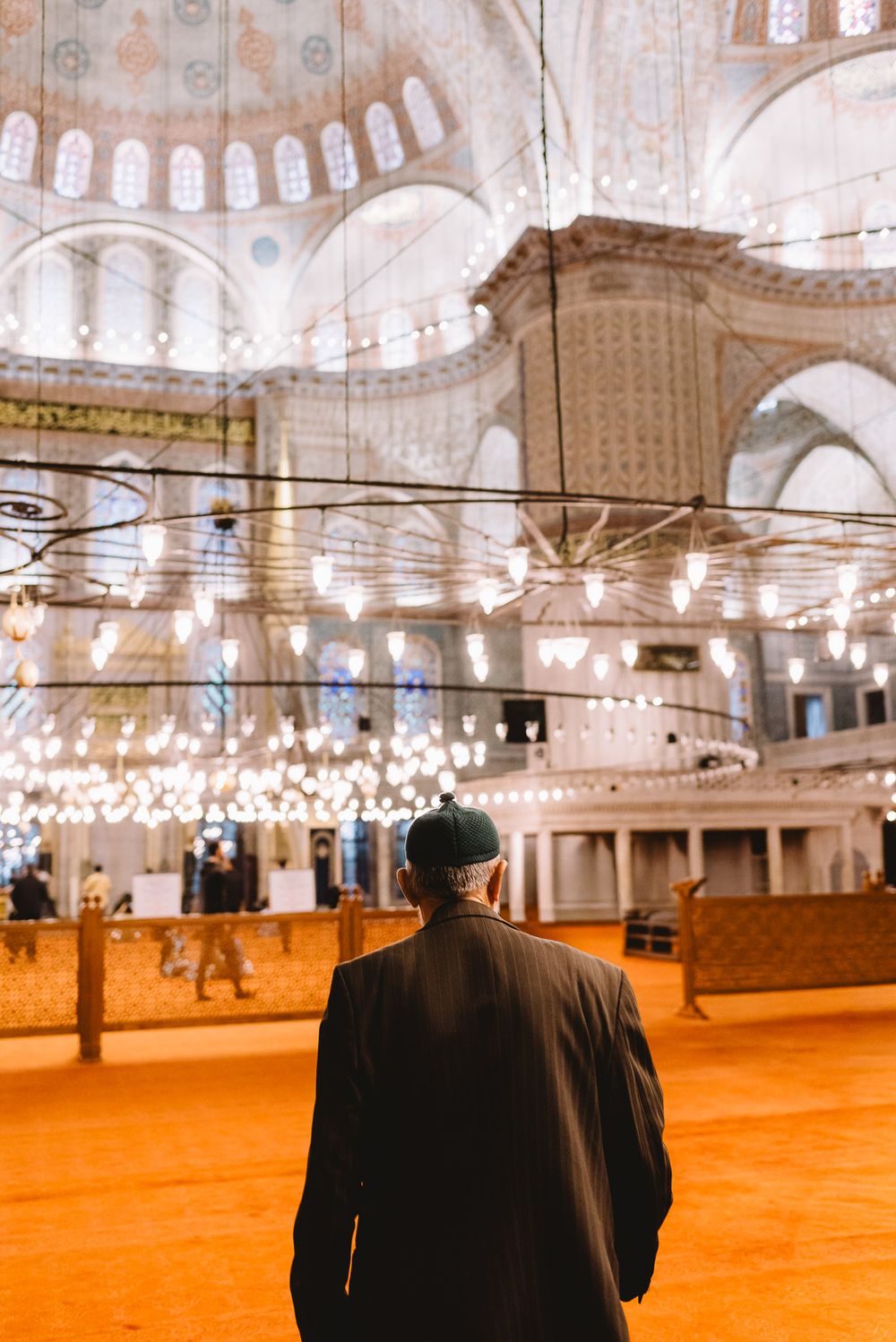 A man in a dark coat stands inside the ornate Blue Mosque in Istanbul, admiring its grand domes and glowing chandeliers.