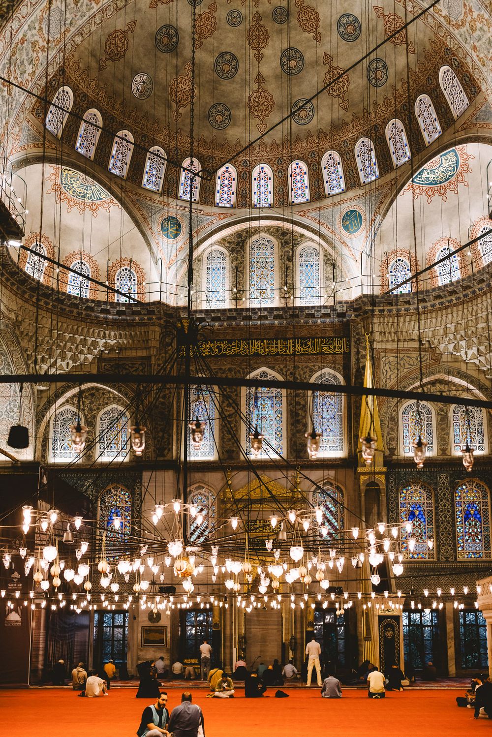 Interior of Blue Mosque Istanbul with ornate domes stained glass windows chandeliers and worshippers praying on red carpet.
