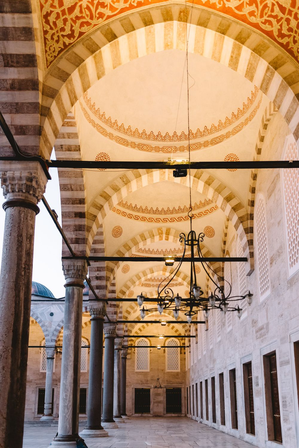 Ornate arched ceiling with intricate Islamic geometric patterns and chandelier in a historic Ottoman mosque courtyard.