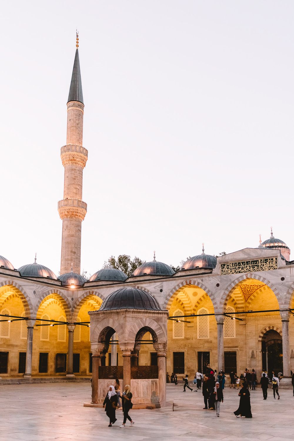 Visitors walk through the illuminated courtyard of Süleymaniye Mosque at dusk, with a tall minaret rising against the sky.