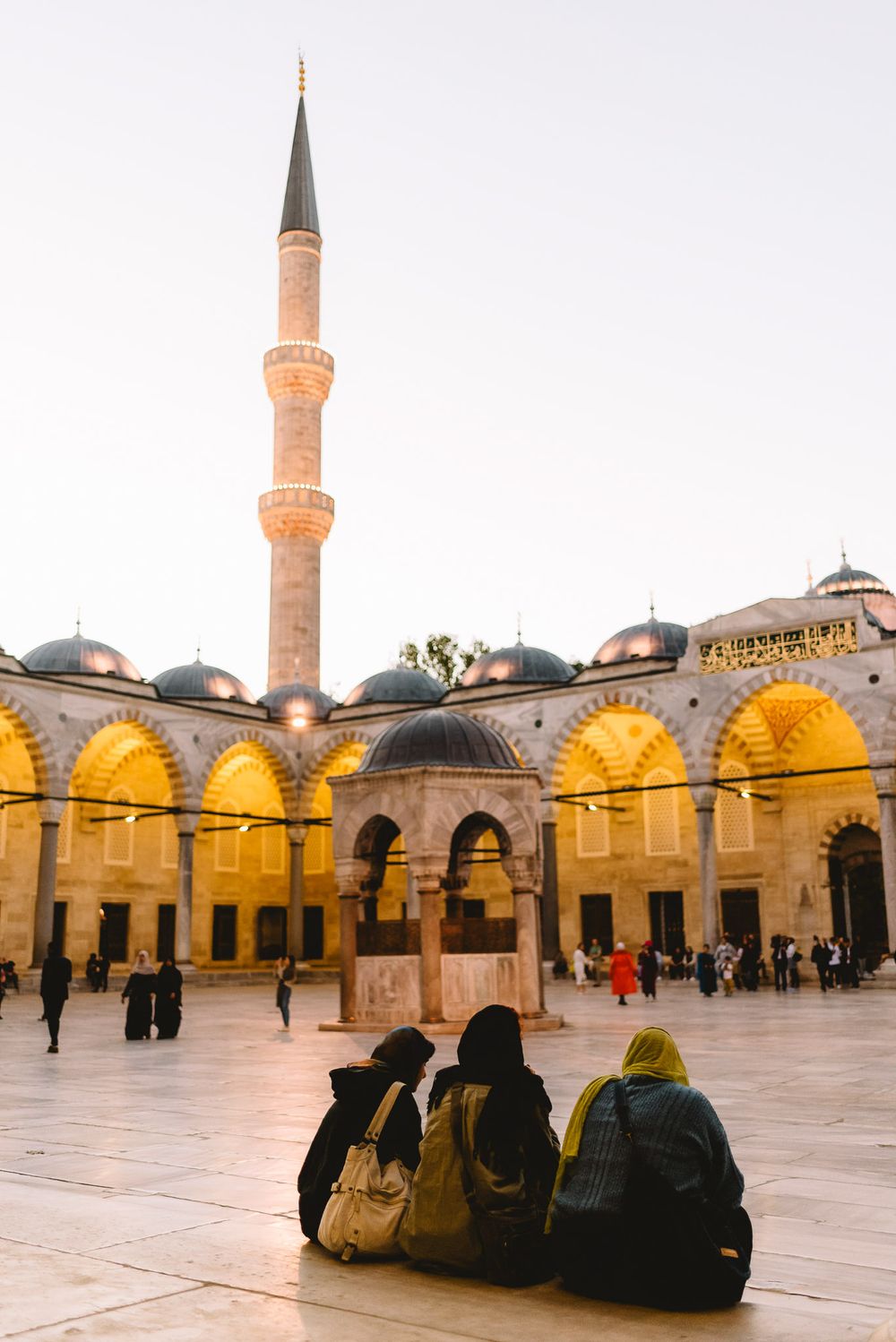 Three women sit in the courtyard of the illuminated Suleymaniye Mosque at dusk, with a minaret rising against the sky.
