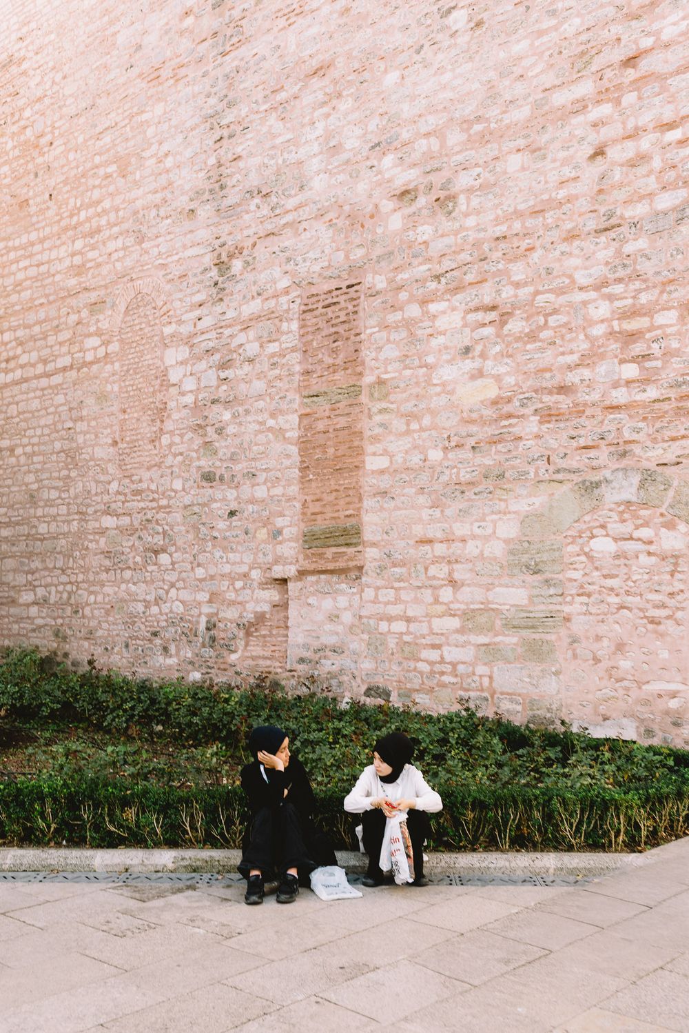 Two people sitting on steps beside a large ancient brick wall with green hedges in a historic courtyard.