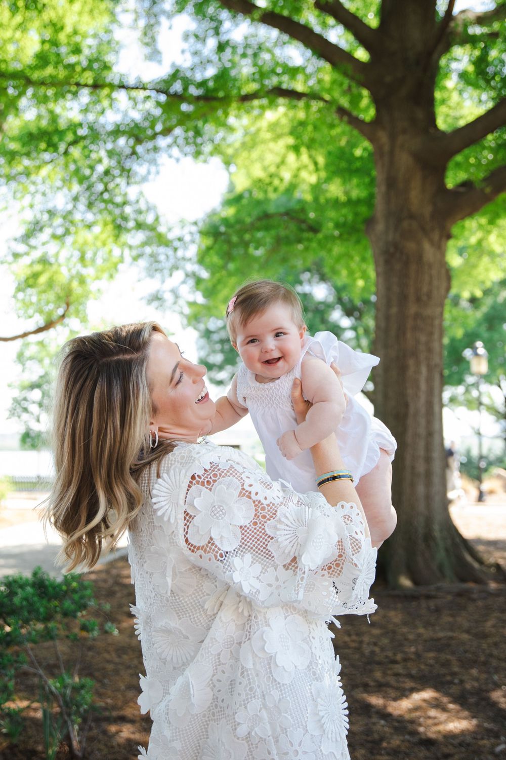 A smiling woman in white lace dress holds a laughing baby girl outdoors under green trees.