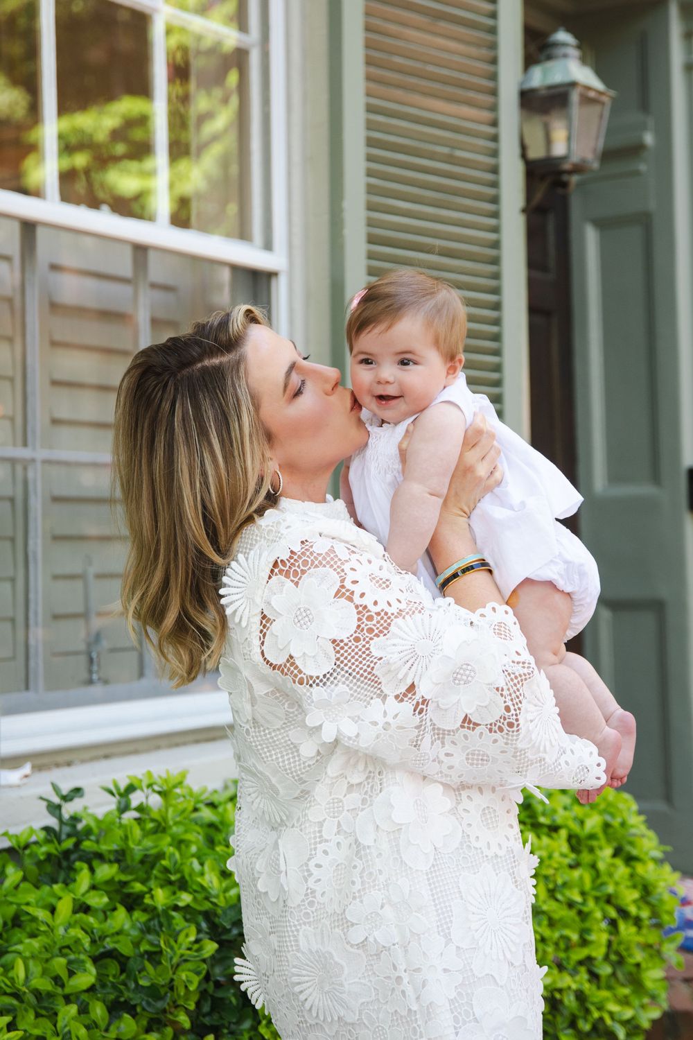 A woman in a white lace dress kisses a smiling baby she holds up outside a home.