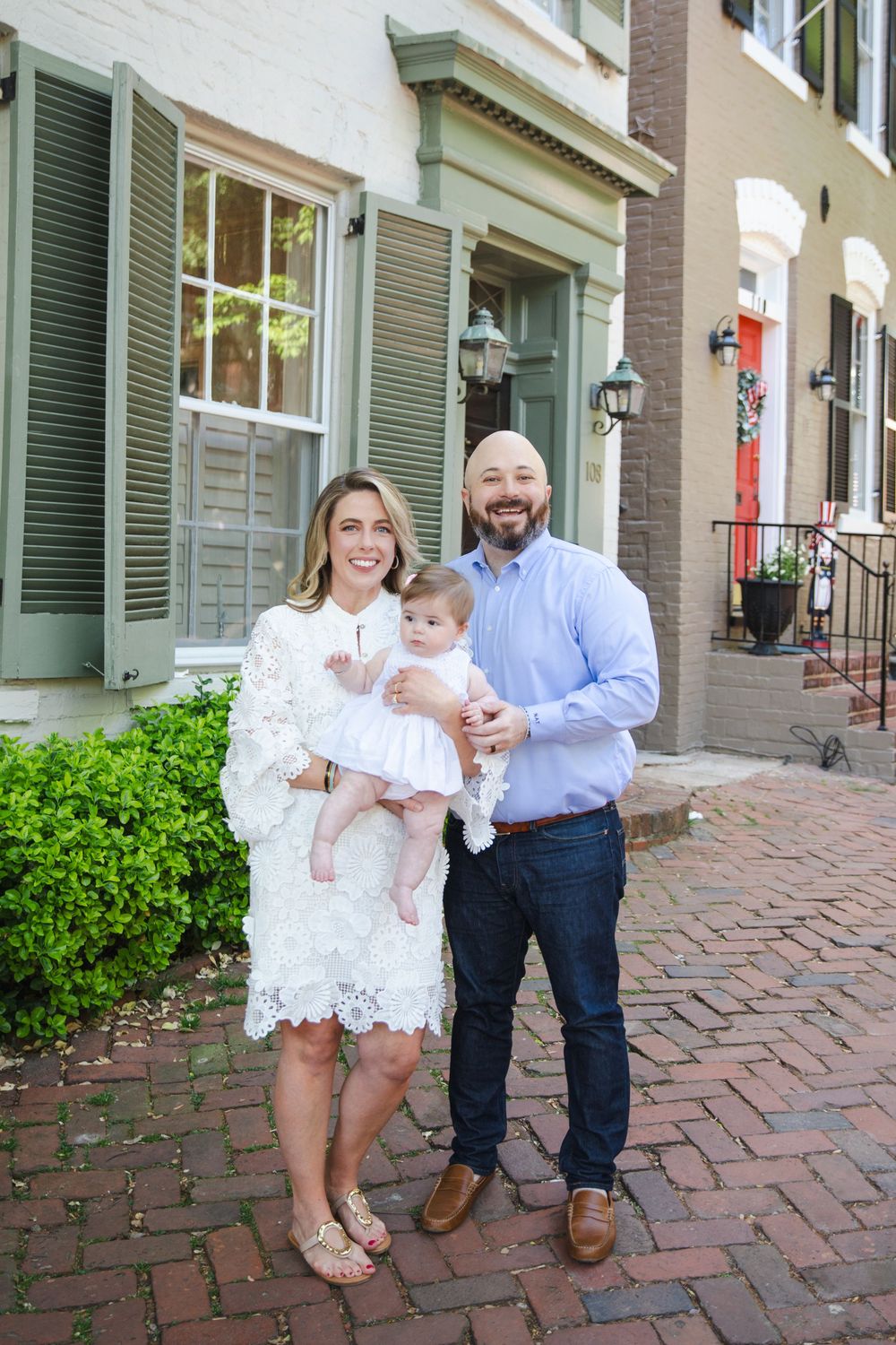 Happy family of three posing on brick sidewalk outside historic townhomes, mother in white lace dress holding baby girl.
