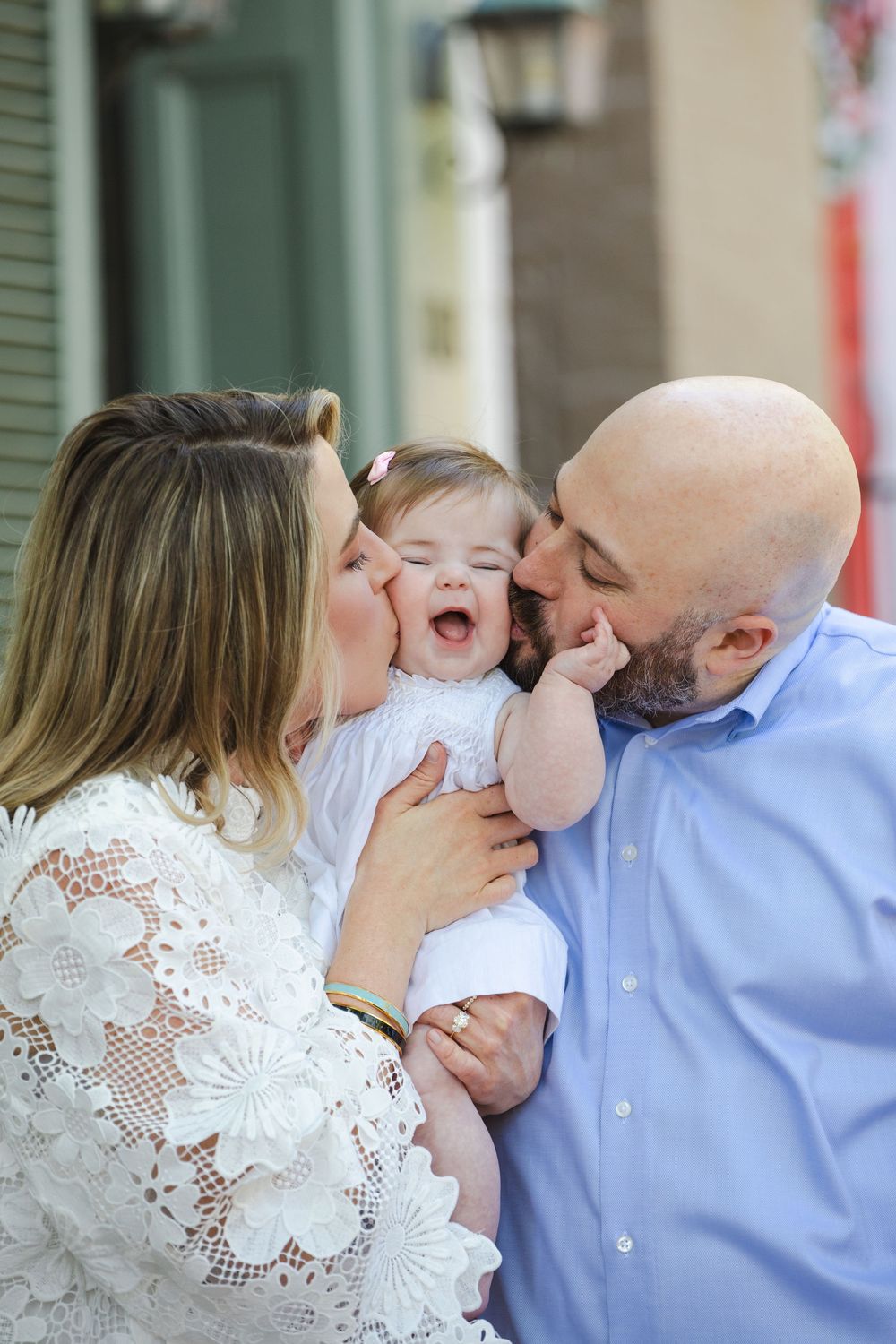 Parents kissing their laughing baby girl on both cheeks outdoors in a heartwarming family portrait.