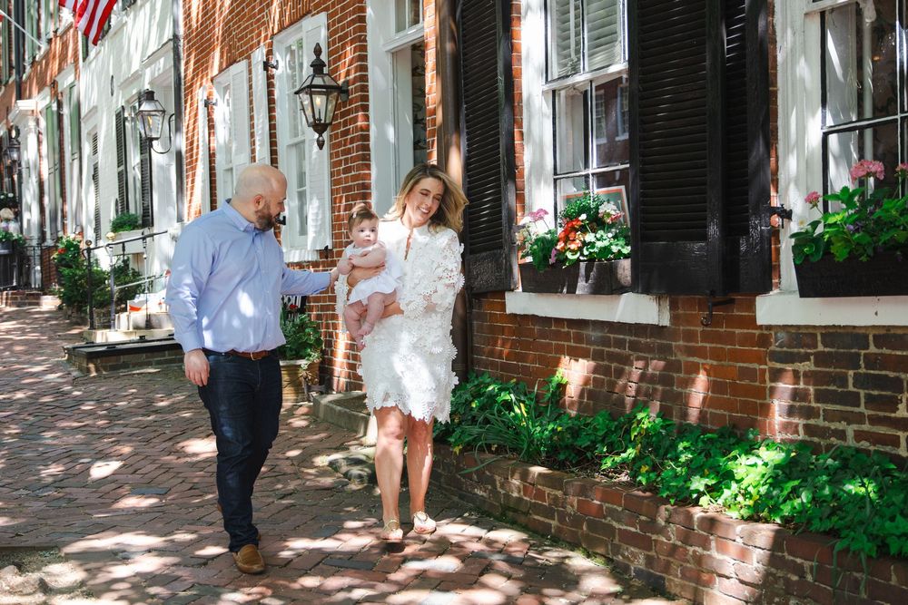 Family of three strolling on a charming brick sidewalk in Old Town Alexandria, parents holding baby girl in white dress.