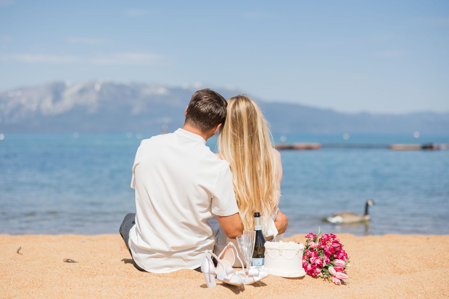 Couple celebrating on a boat with champagne, snow-capped mountains and calm blue lake in the background.