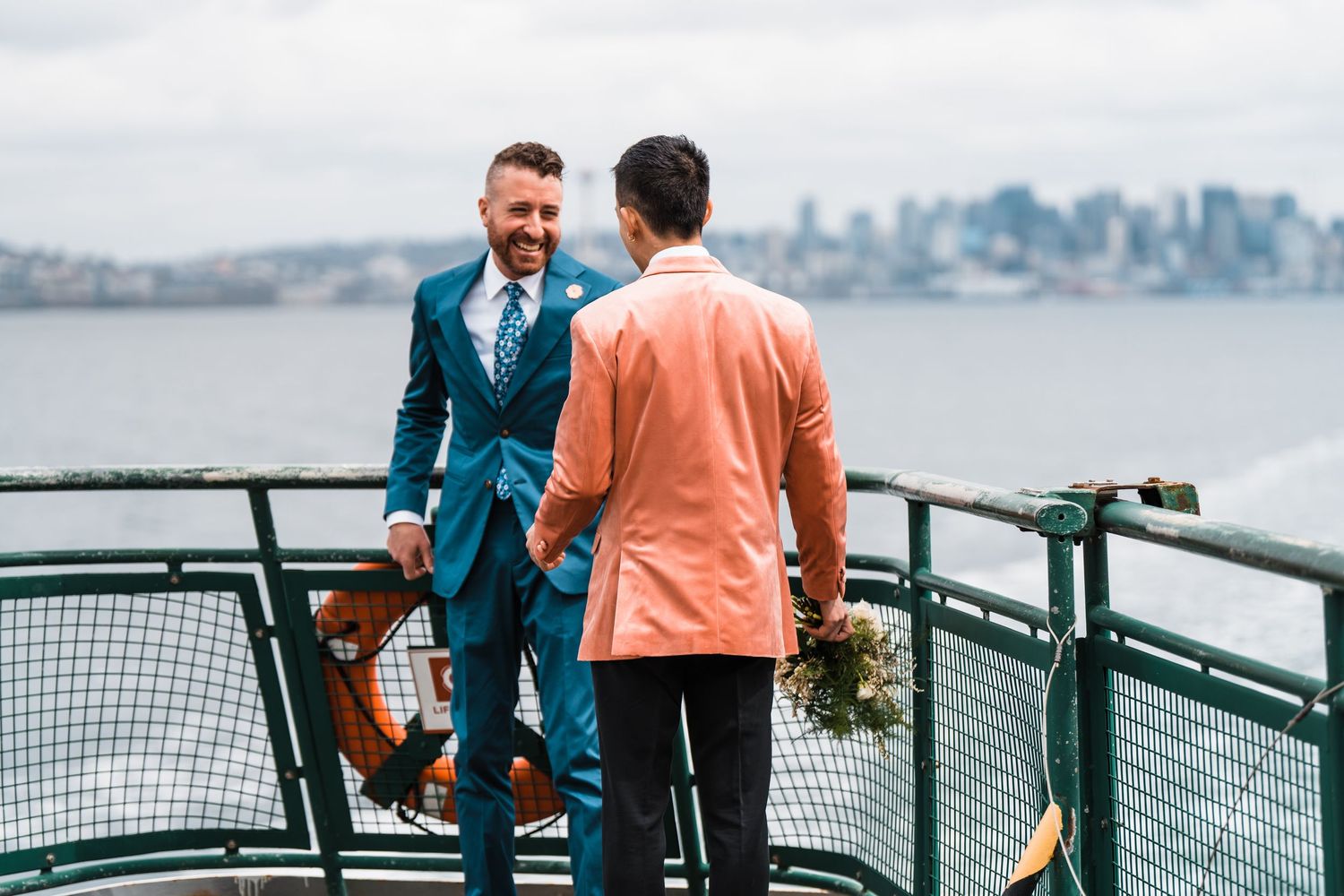 Two grooms in blue and salmon suits hold hands on a ferry with Seattle skyline backdrop. Bainbridge island ferry elopement