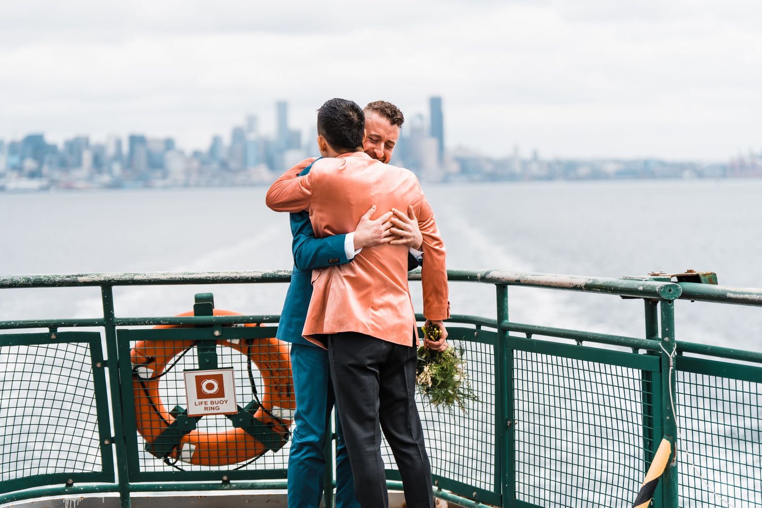 Two people embrace on a ferry deck with Seattle skyline and Puget Sound in the background. Bainbridge island ferry elopement