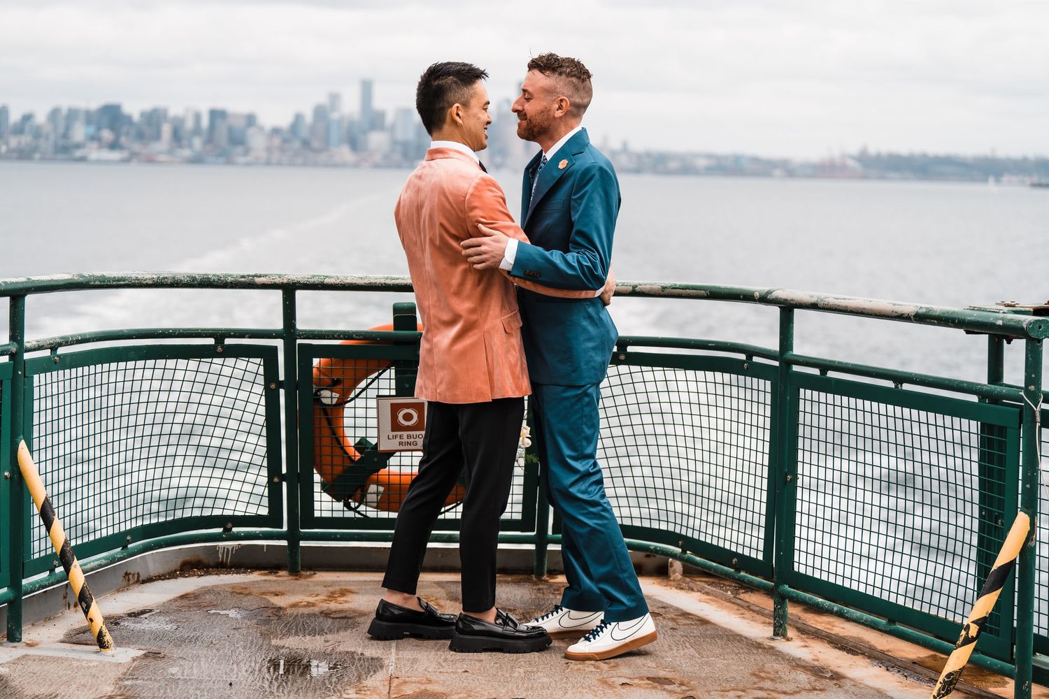 Two grooms in teal and salmon suits embrace on a ferry deck with city skyline backdrop. Bainbridge island ferry elopement