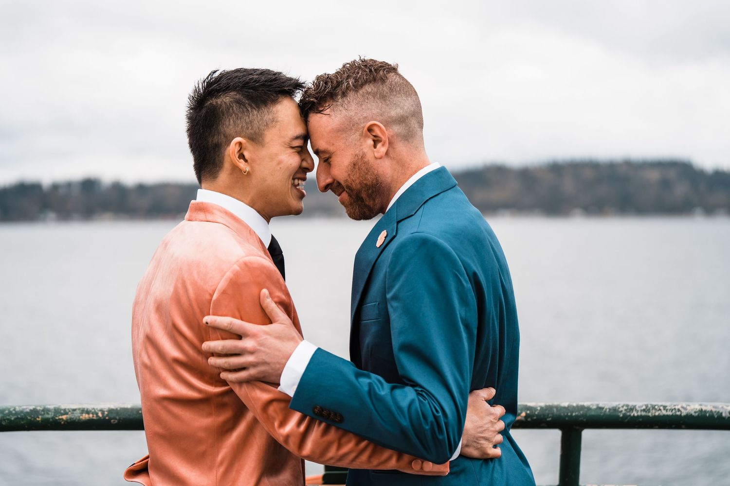 Two grooms in coral and teal suits share a romantic moment by a waterfront railing on their wedding day. Bainbridge island ferry elopement