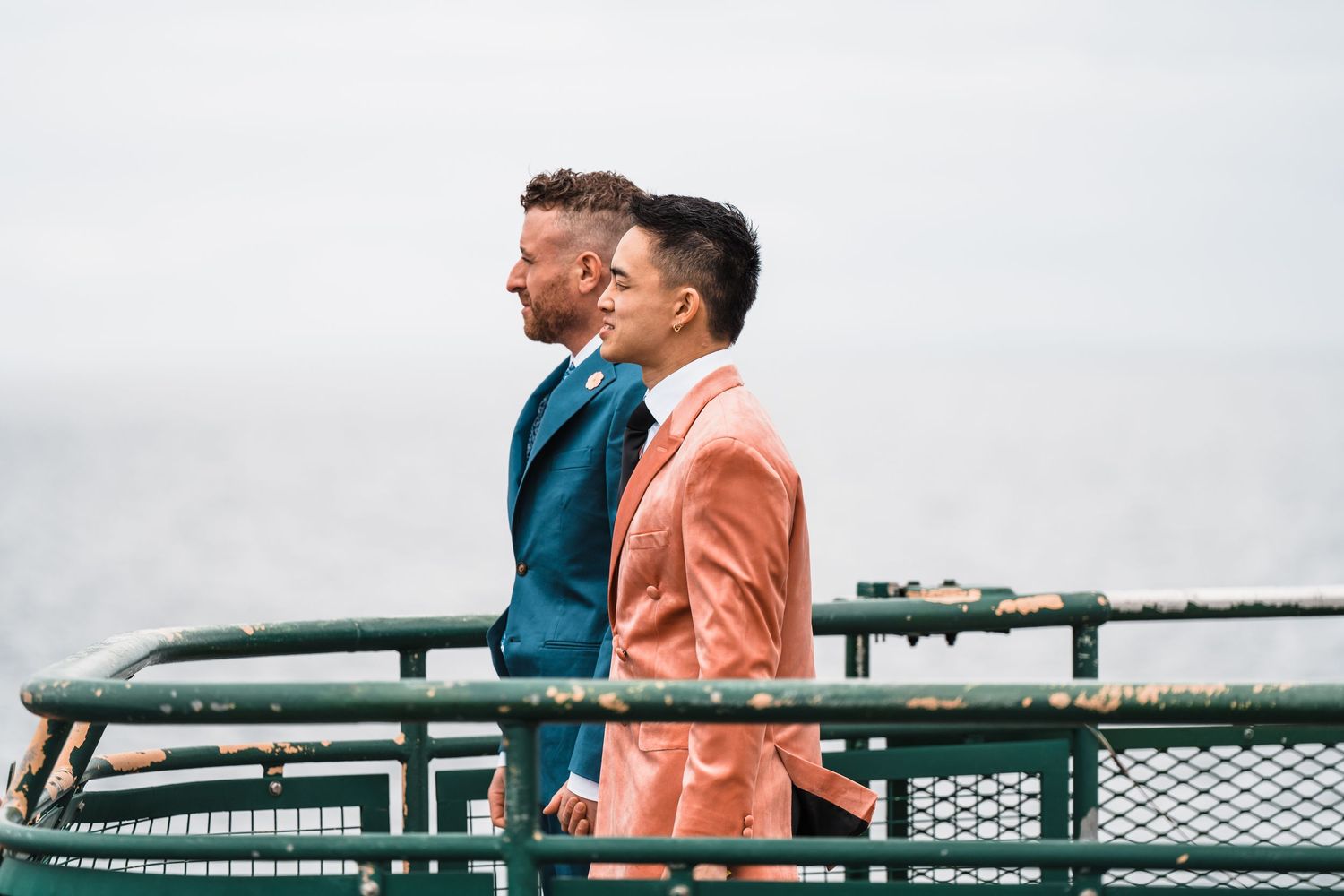 Two grooms in colorful suits, one teal and one coral, stand together on a railing overlooking a cloudy sky. Bainbridge island ferry elopemen