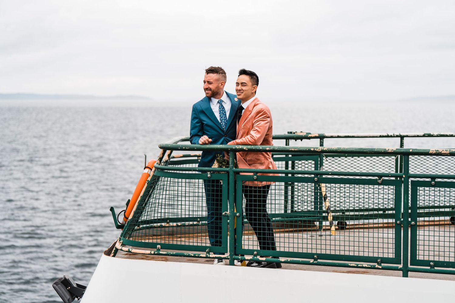 Two men in suits stand on a ferry deck overlooking the ocean on a cloudy day. Bainbridge island ferry elopement