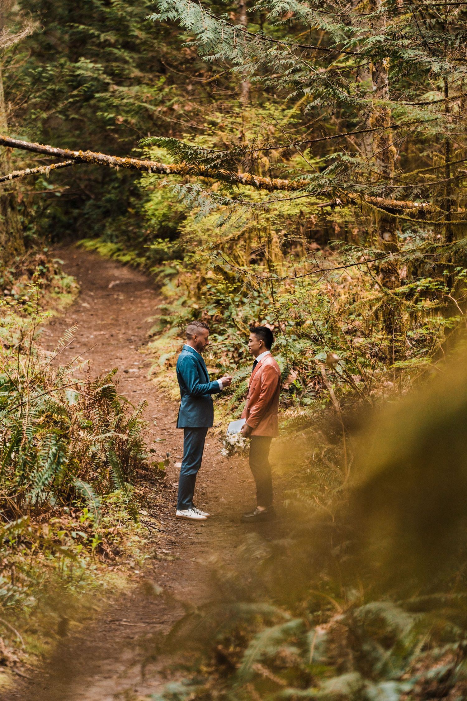 Two grooms exchange vows on a forest trail surrounded by lush green ferns and towering trees during an outdoor wedding. Bainbridge island fe