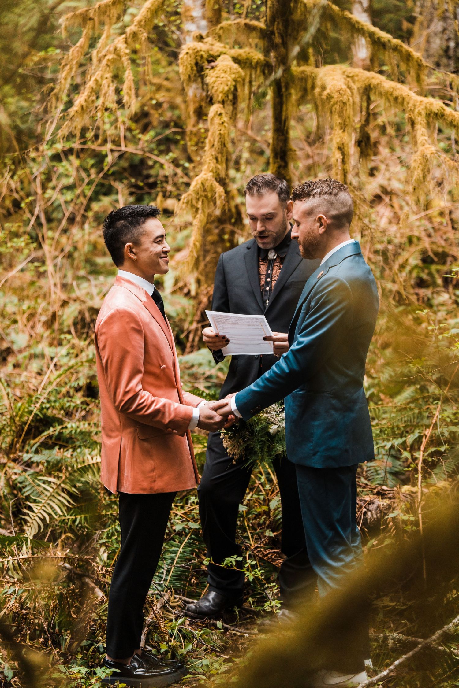 Two grooms hold hands during an outdoor forest wedding ceremony with an officiant reading vows among lush green ferns.Bainbridge island ferr