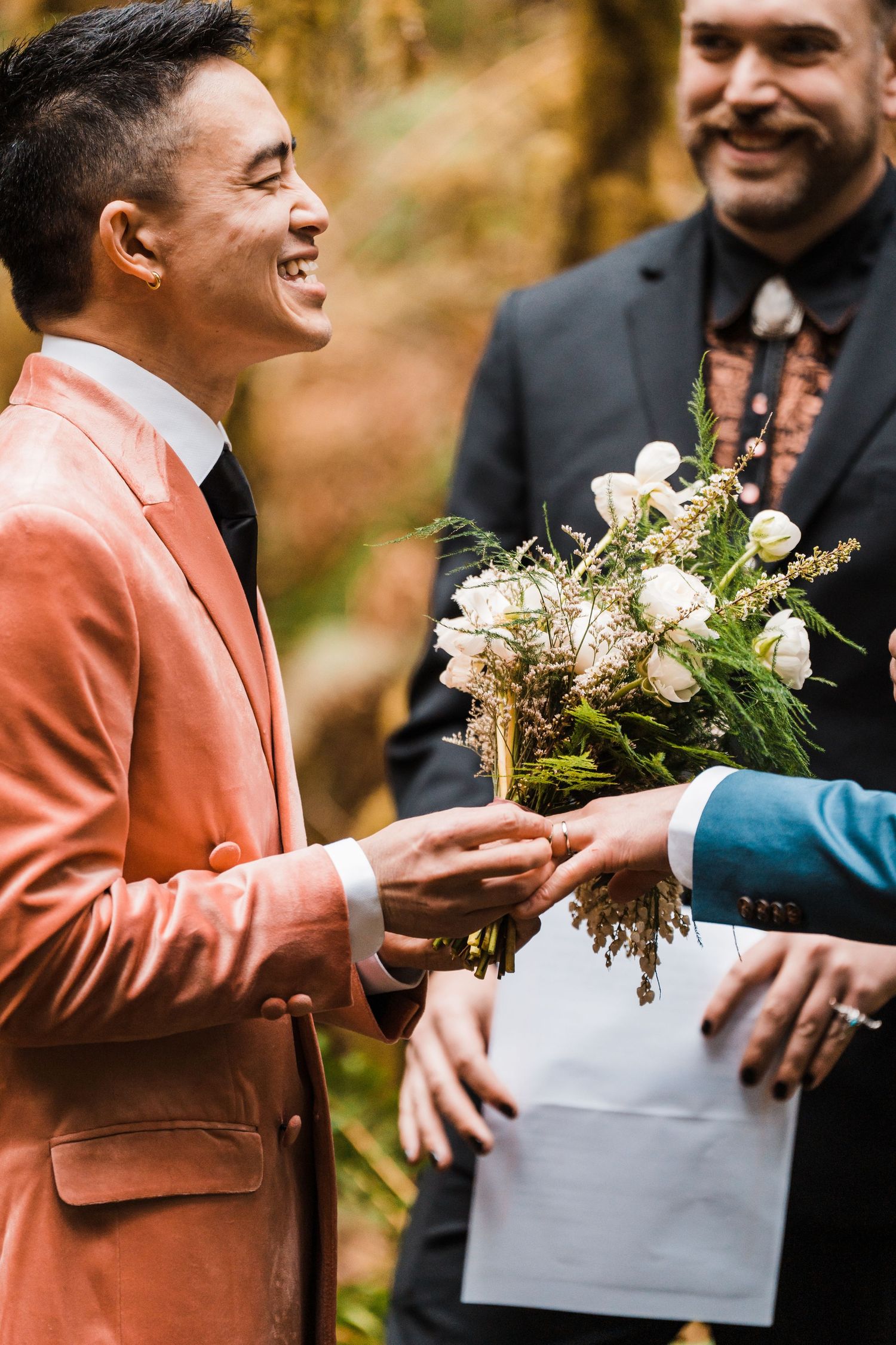 Two grooms smile during outdoor wedding ceremony as one receives white rose bouquet in forest setting. Bainbridge island ferry elopement