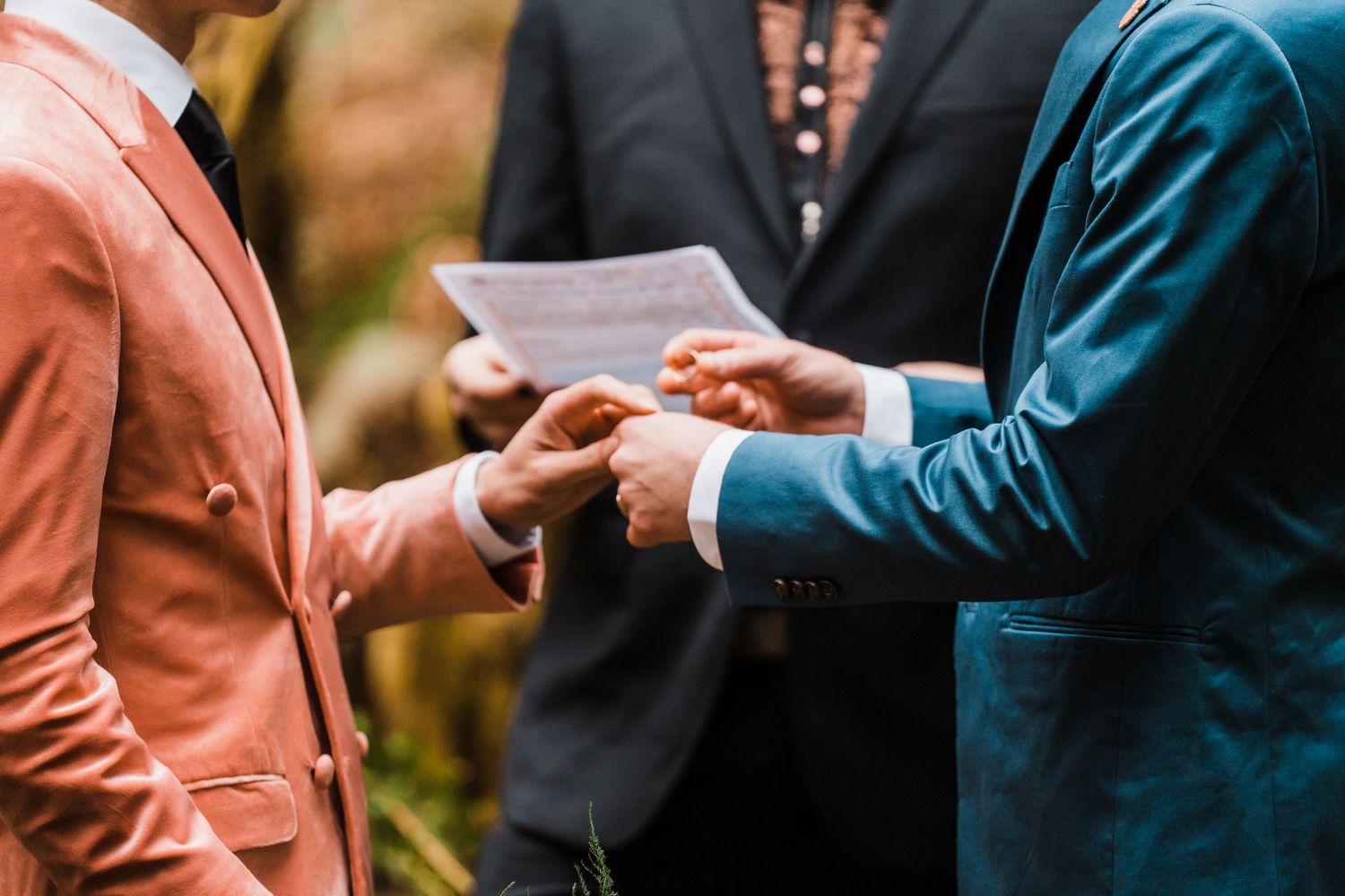Grooms exchange rings during outdoor wedding ceremony, officiant reading vows in background. Bainbridge island ferry elopement