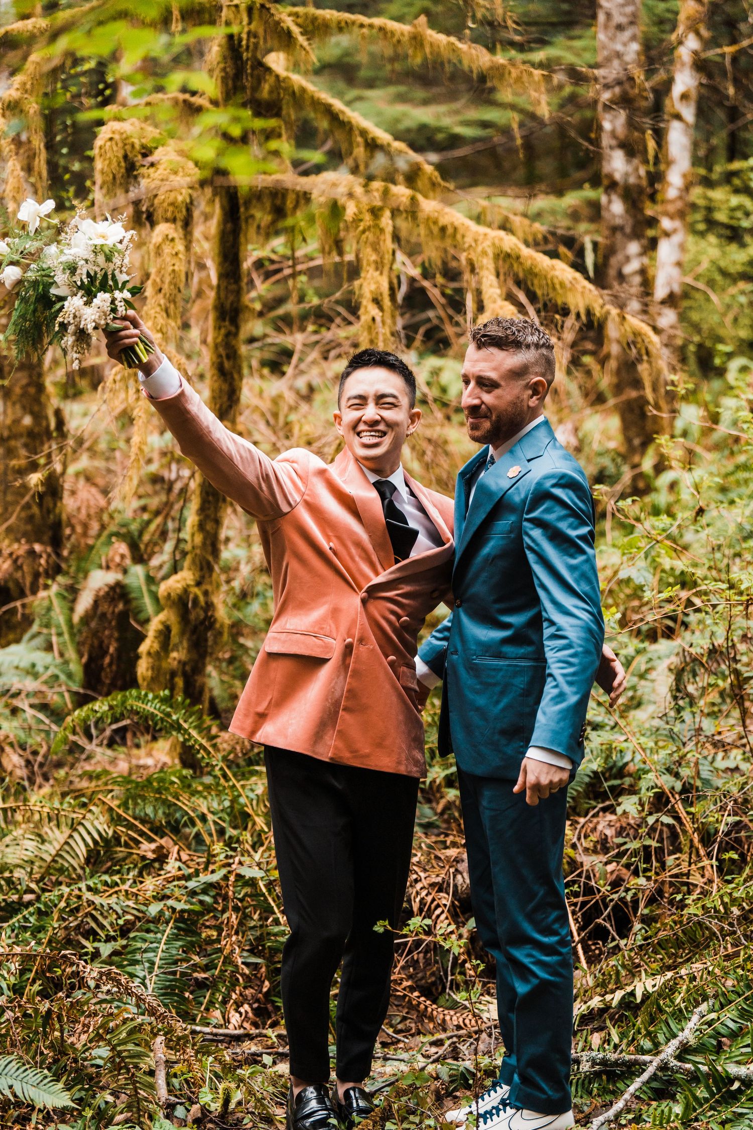 Two grooms in colorful suits celebrate in a lush forest, one raising a white bouquet joyfully. Bainbridge island ferry elopement