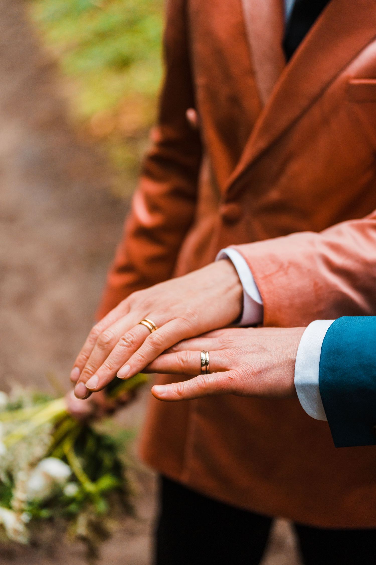 Two grooms displaying gold wedding rings on their hands, one wearing a brown velvet blazer, holding a white floral bouquet.Bainbridge island