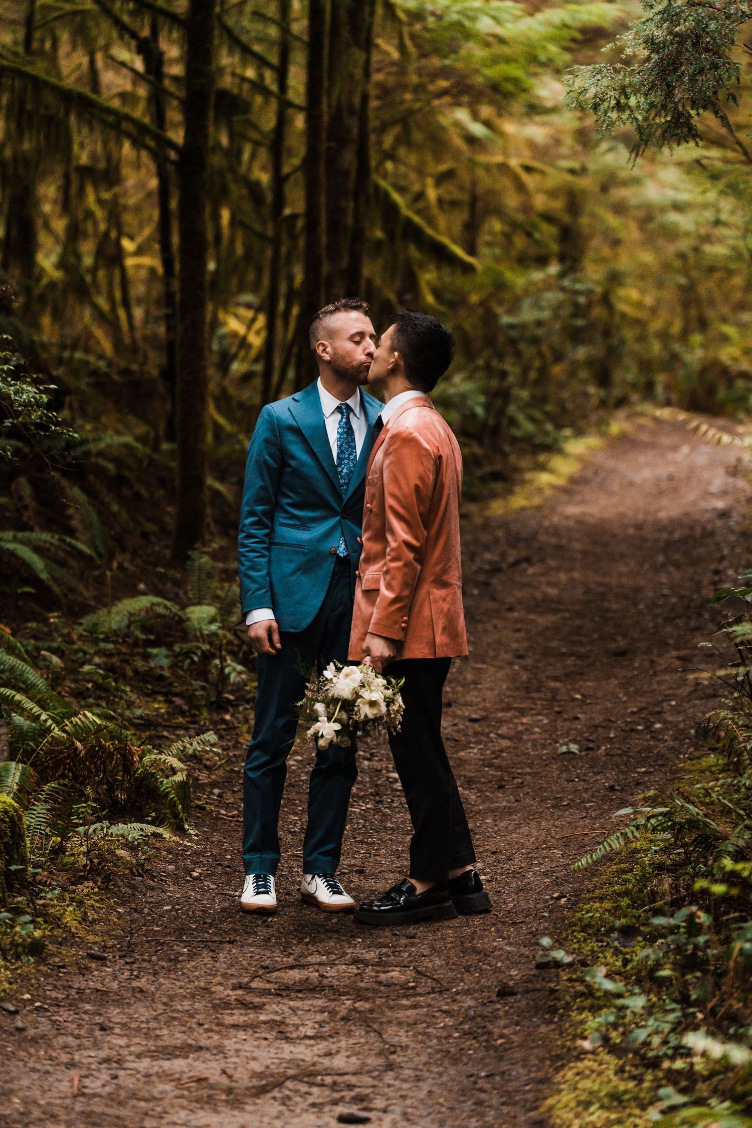 Two grooms share a first kiss on a forest trail, one in a teal suit and one in a rust blazer holding a white bouquet. Bainbridge island ferr