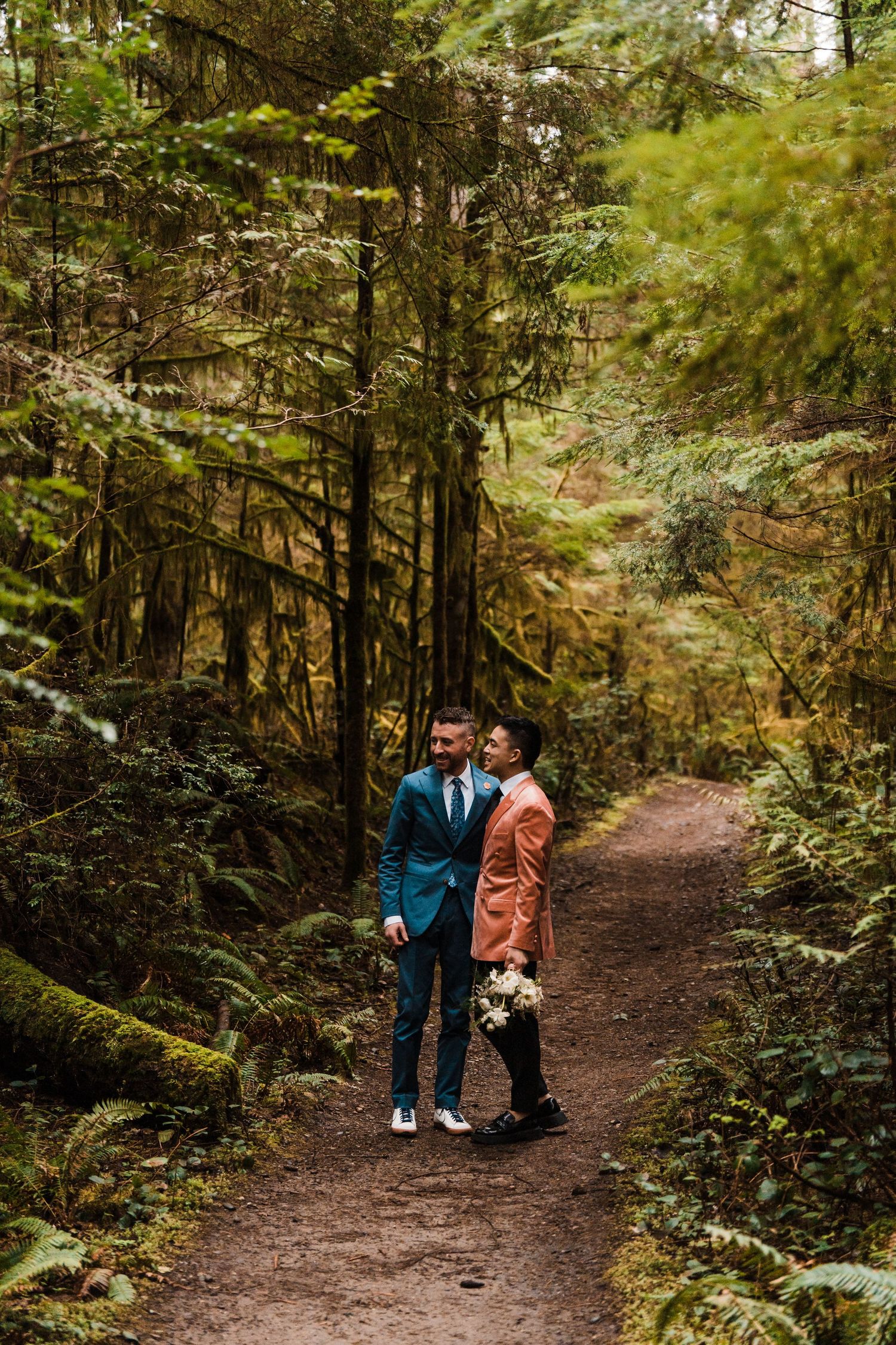 Two grooms in teal and salmon suits stand on a lush forest trail holding a wedding bouquet. Bainbridge island ferry elopement