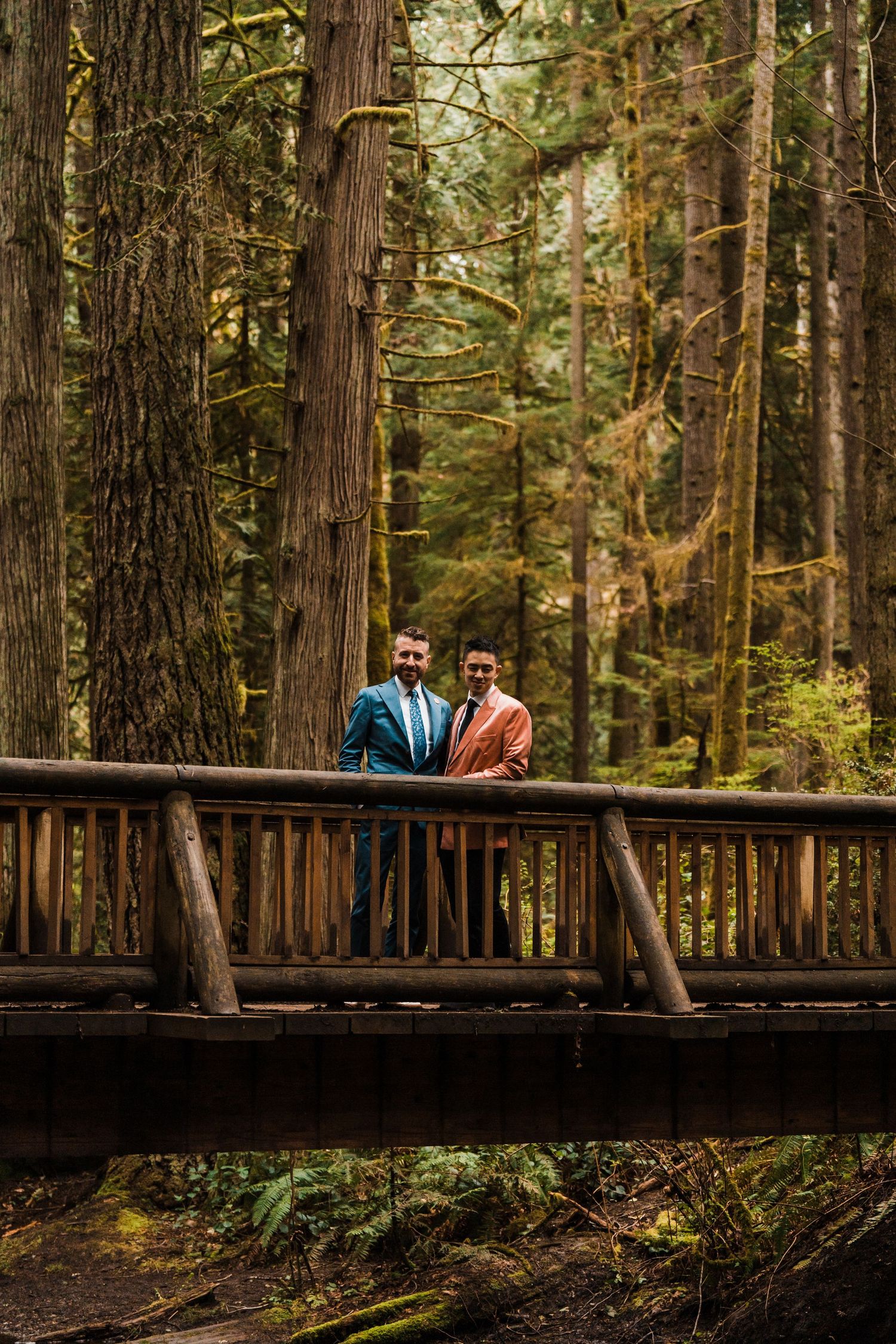 Two grooms in blue and salmon suits pose together on rustic wooden bridge surrounded by tall forest trees. Bainbridge island ferry elopement