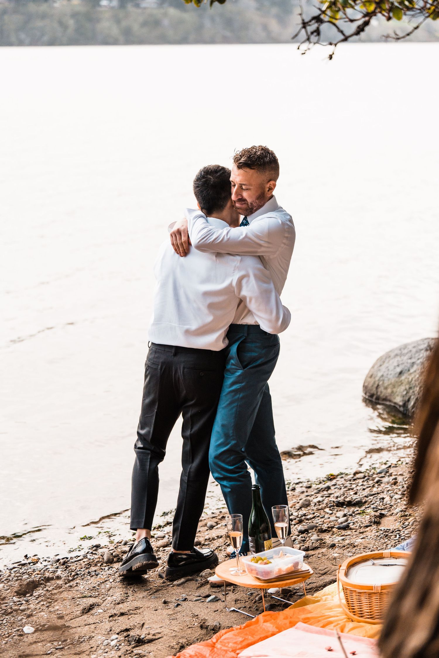 Two grooms share a heartfelt embrace by a sunlit lakeside during their outdoor wedding celebration. Bainbridge island ferry elopement