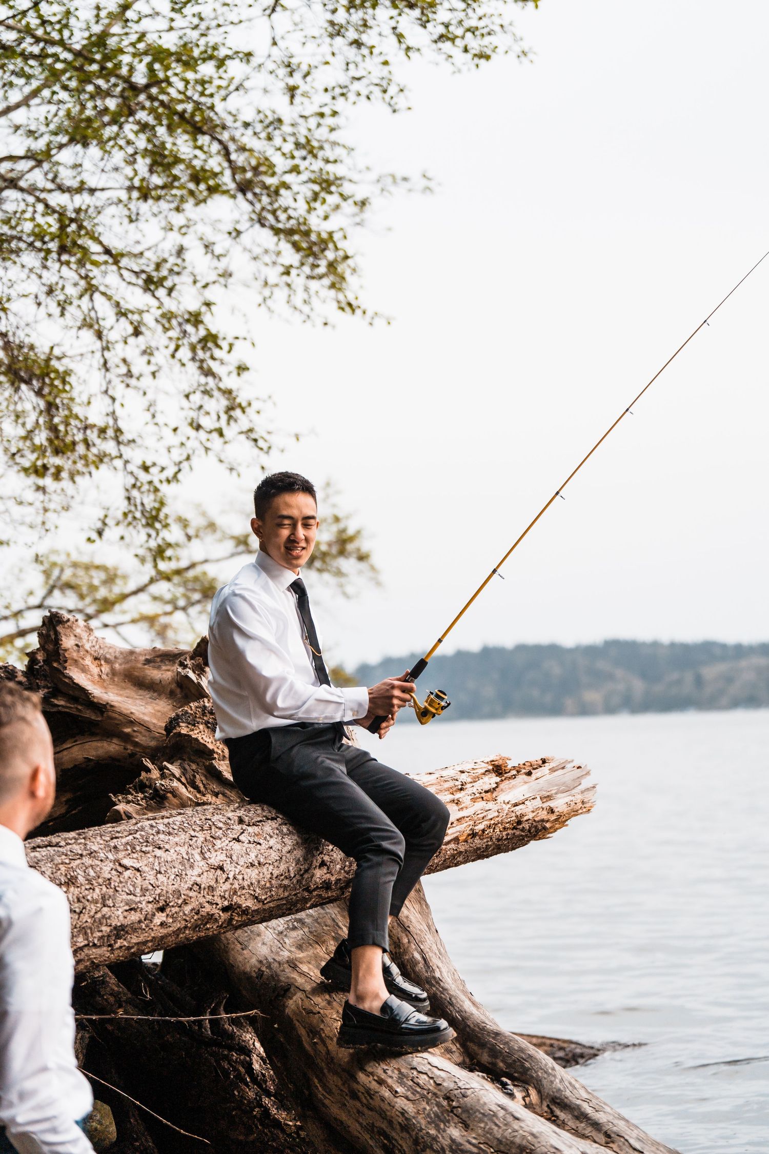 A groom in a white shirt and tie sits on driftwood fishing by a calm lake on his wedding day. Bainbridge island ferry elopement