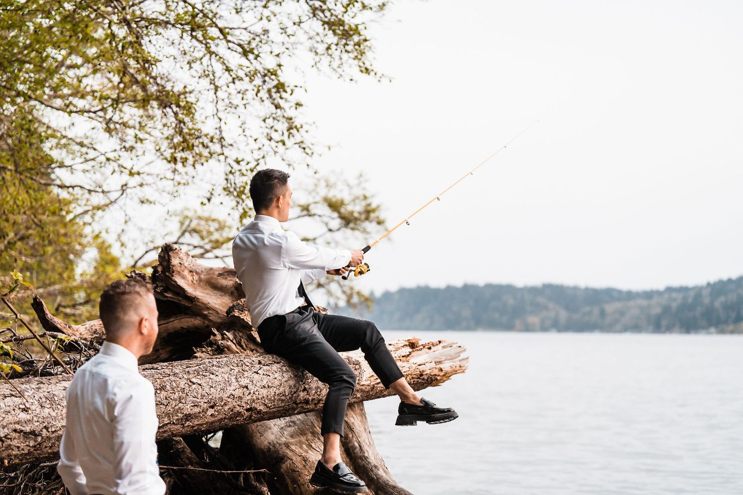 Two men relaxing by a lake, one fishing from a pile of logs near the water's edge surrounded by trees. Bainbridge island ferry elopement