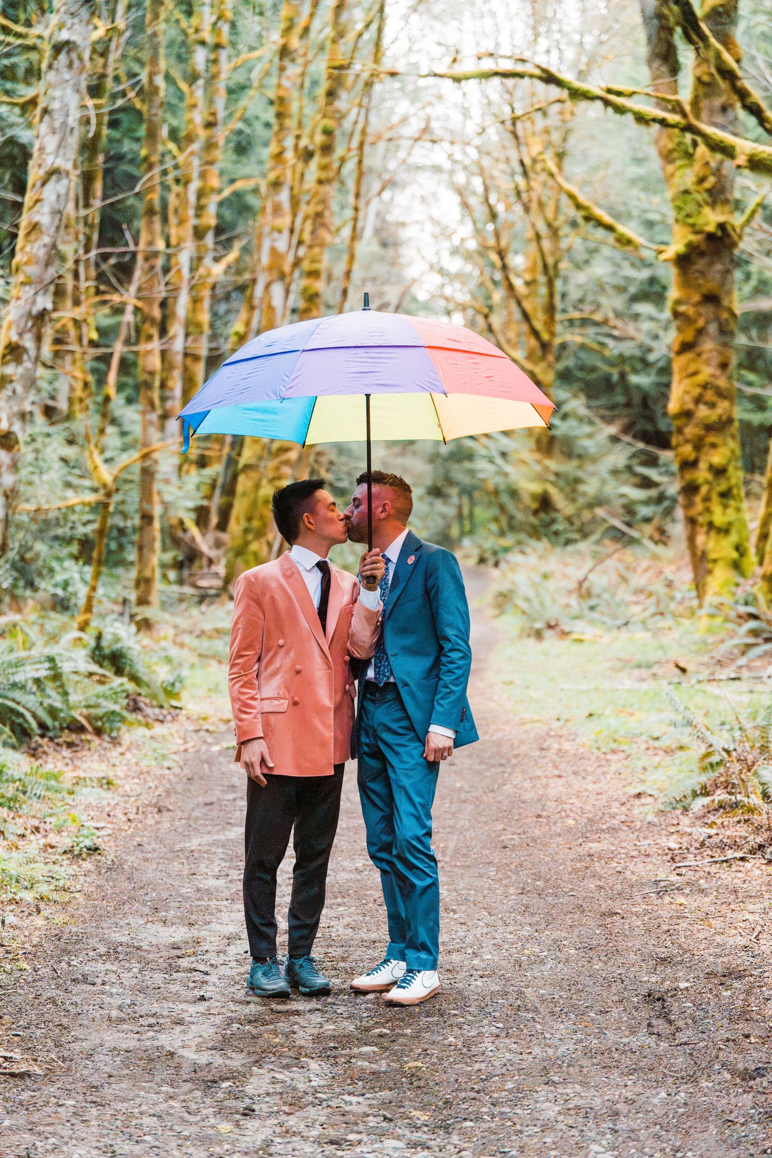 Two grooms share a kiss under a rainbow umbrella on a forest path, one in pink and one in teal suit.Bainbridge island ferry elopement
