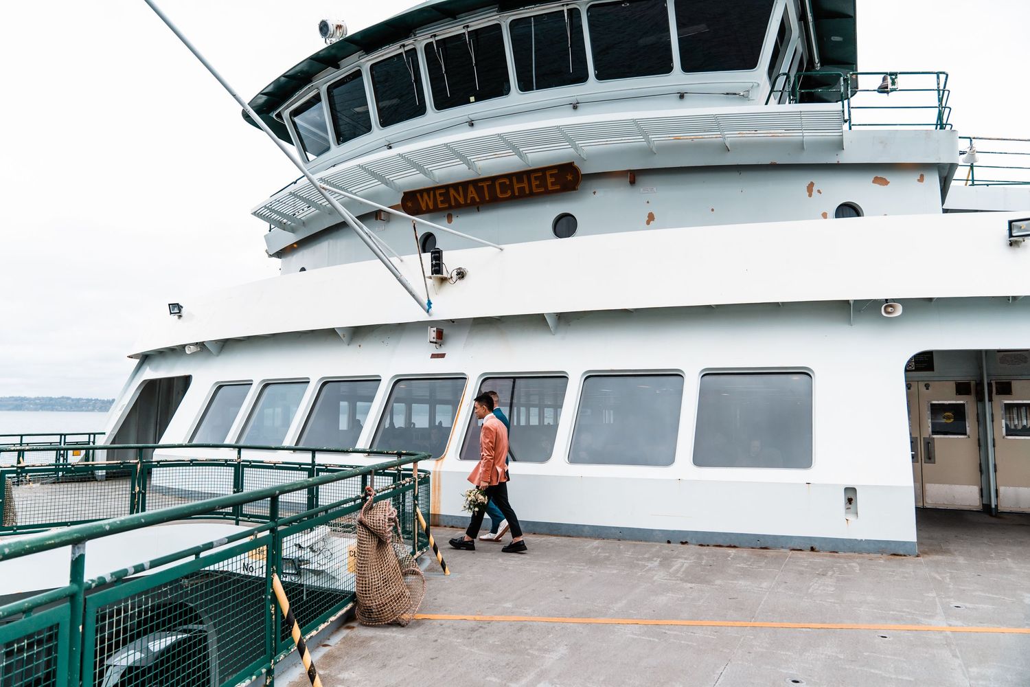 A person in an orange jacket walks along the exterior deck of a large white ferry boat.Bainbridge island ferry elopement