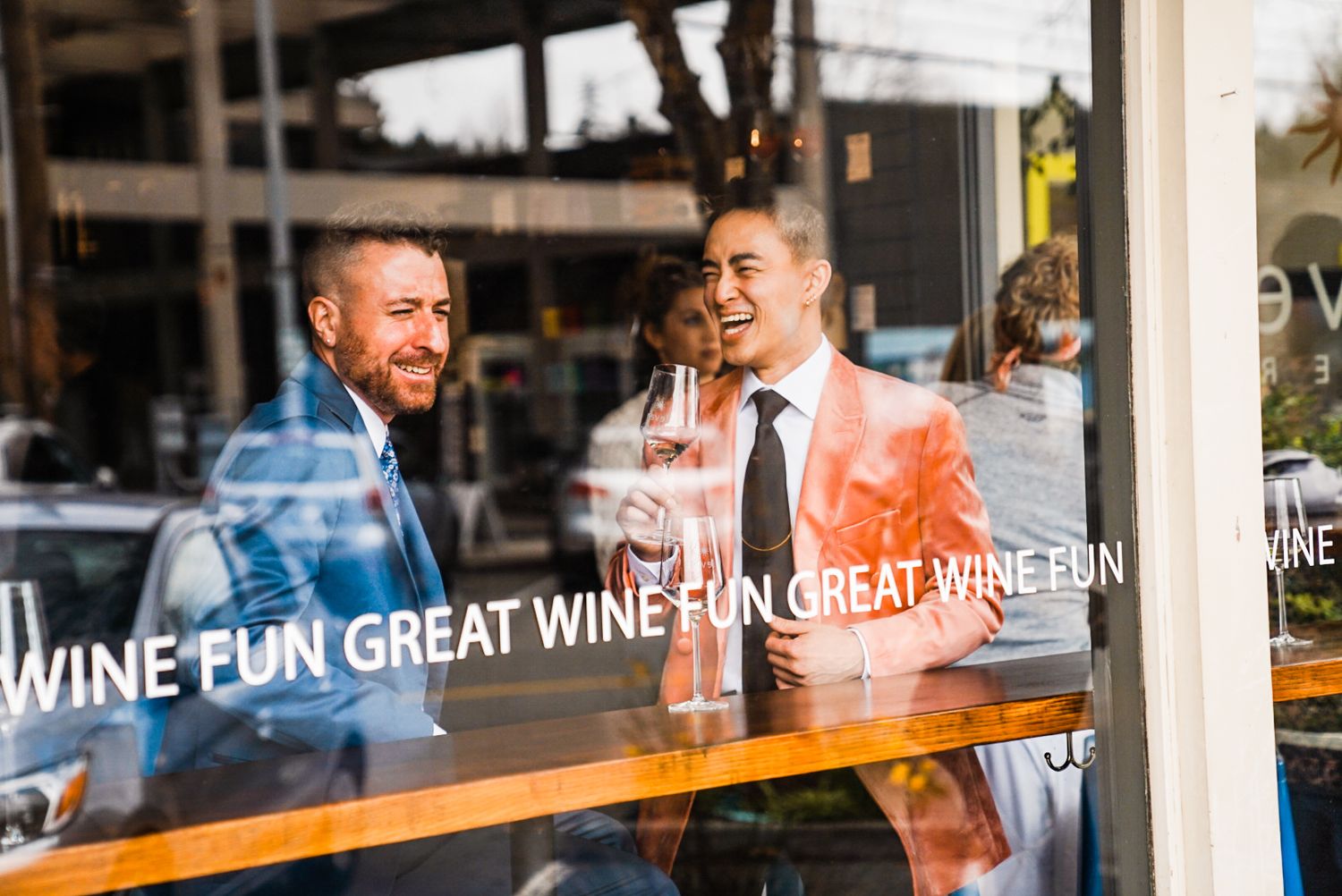 Two men in suits laughing together inside a wine bar with 'Great Wine Fun' text on the window.Bainbridge island ferry elopement