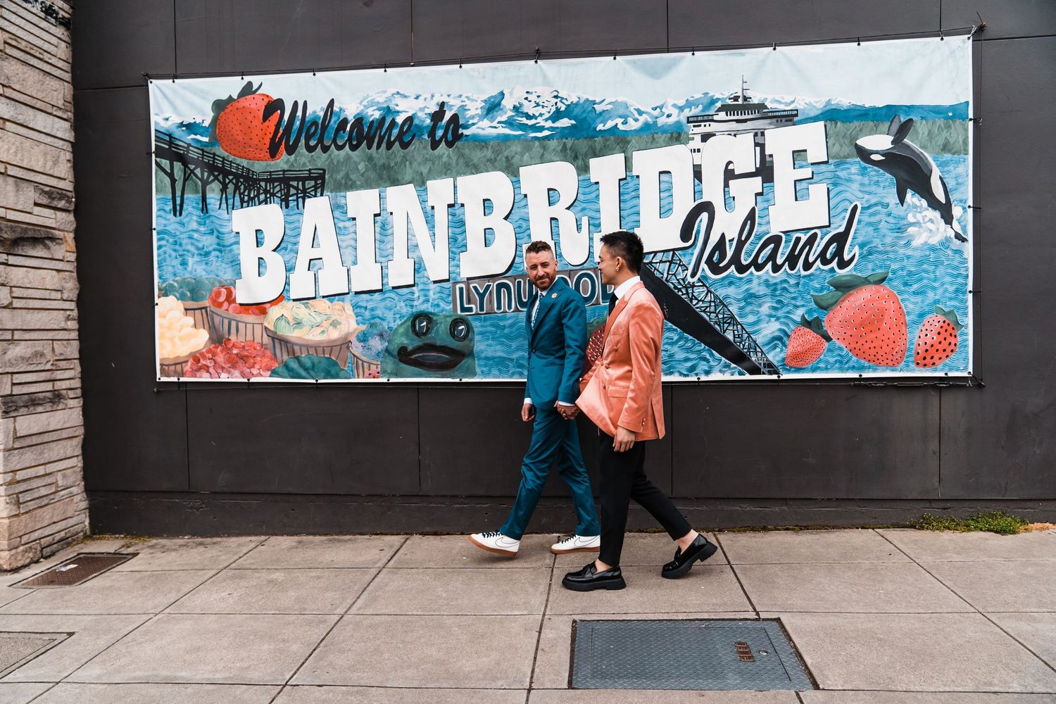 A couple walks past a colorful Welcome to Bainbridge Island mural sign on a sunny day.Bainbridge island ferry elopement