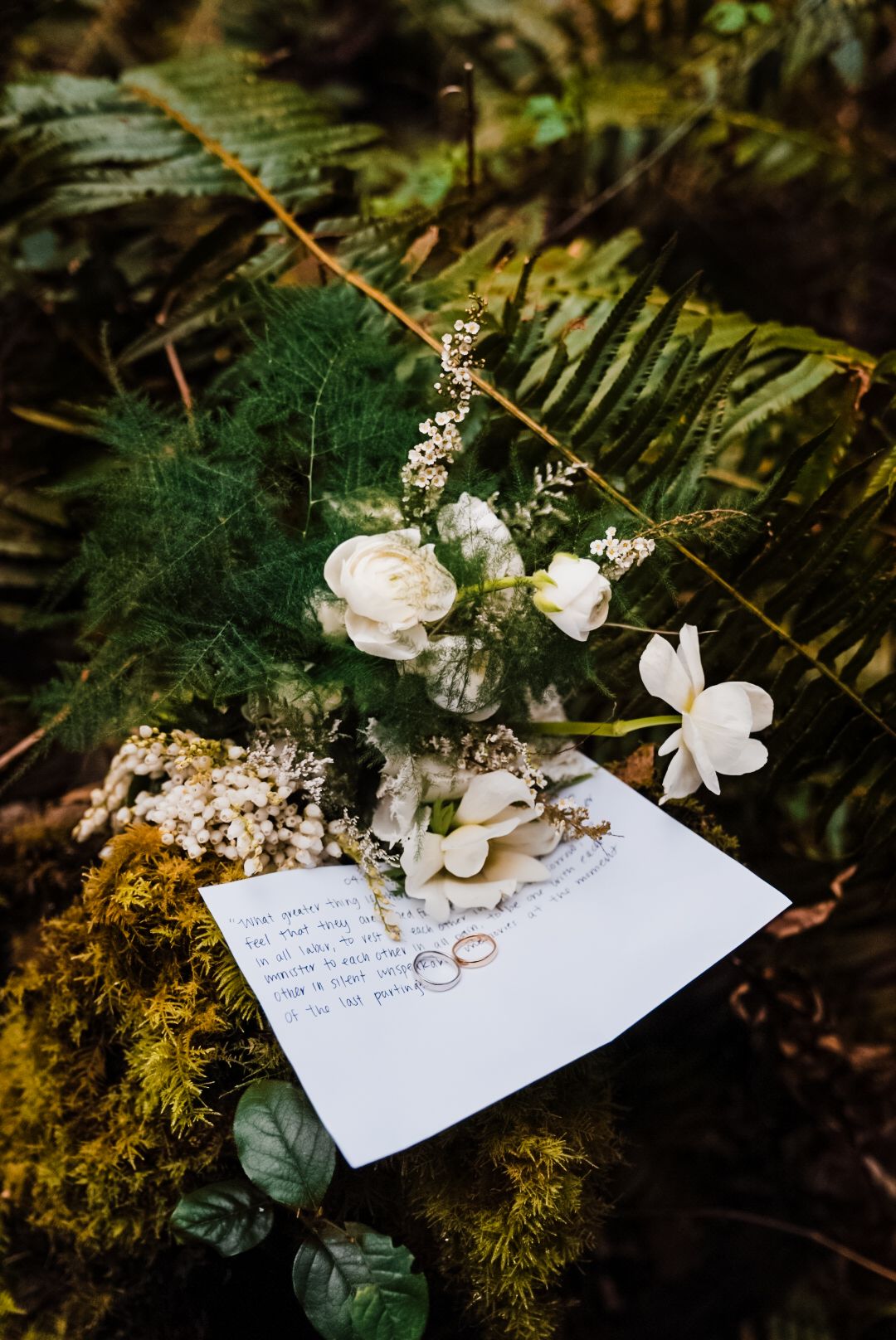 Wedding rings rest on handwritten vows beside white roses and ferns on mossy forest ground. Bainbridge island ferry elopement