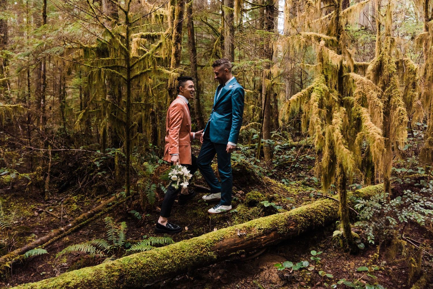 Two grooms in colorful suits walk hand-in-hand on a mossy log in a lush, green forest wedding elopement.Bainbridge island ferry elopement