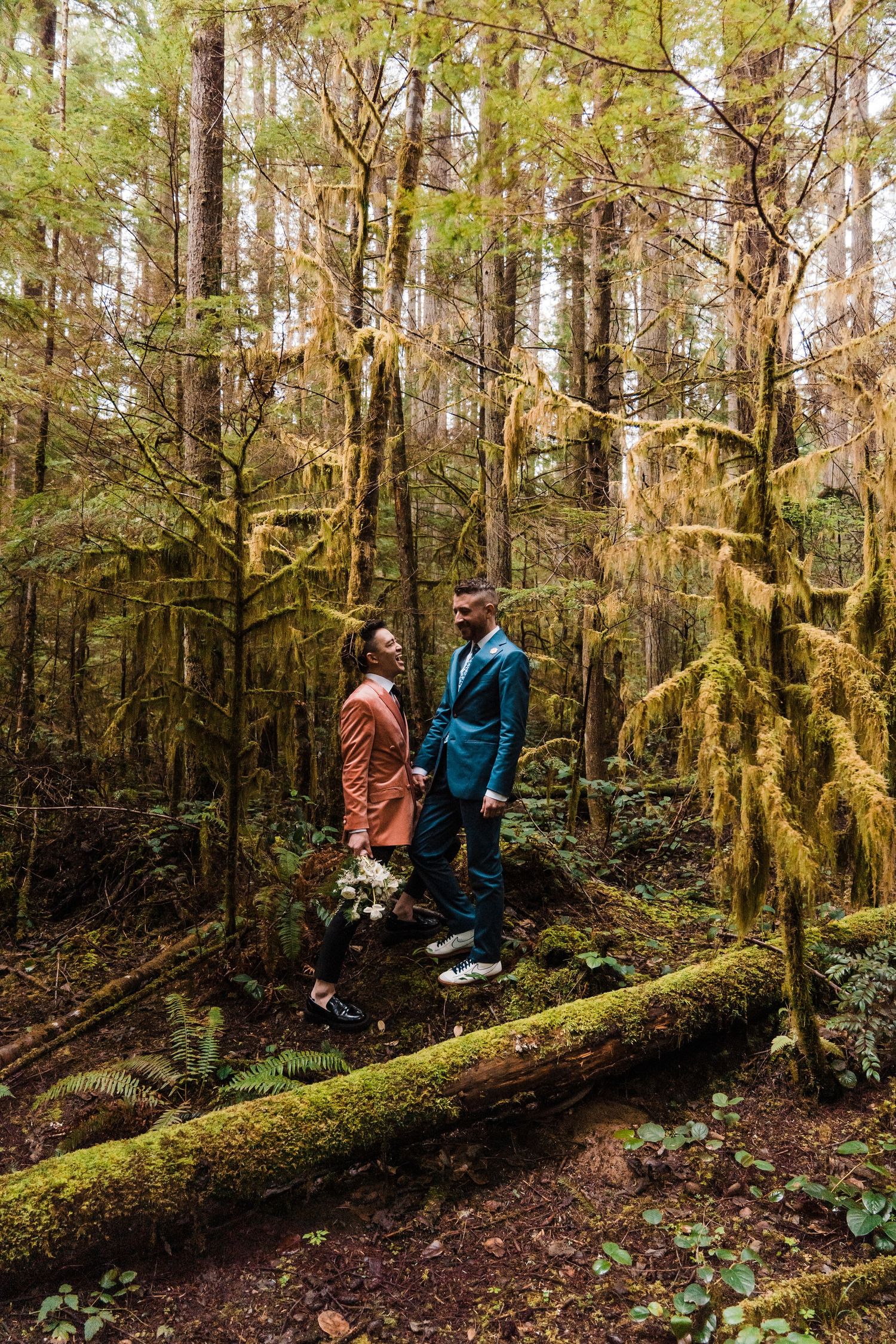 Couple in coordinating rust and teal suits stand on a mossy log in a lush Pacific Northwest forest. Bainbridge island ferry elopement
