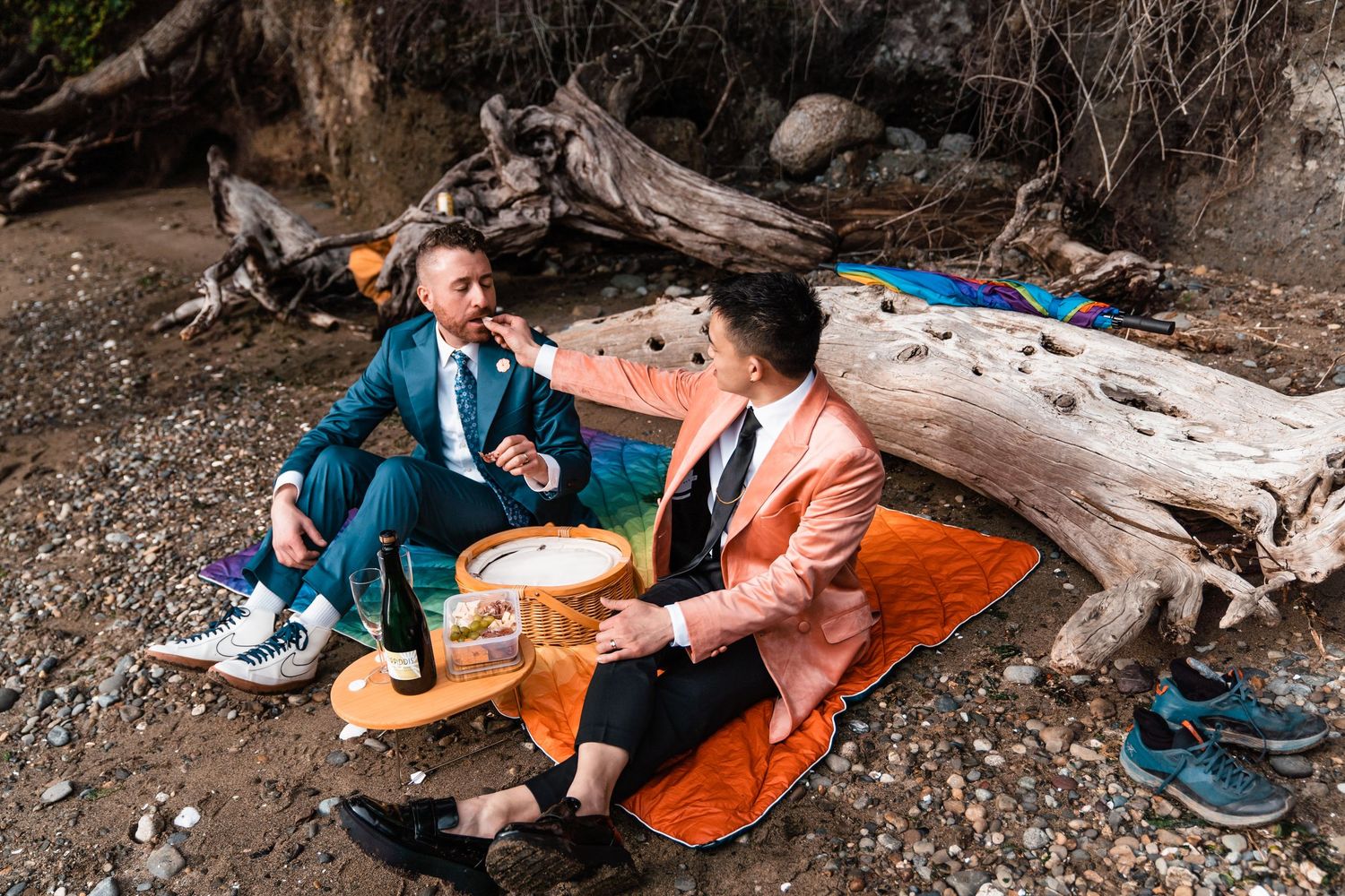 Two men in colorful blazers enjoy a romantic outdoor picnic near driftwood on a rocky beach. queer elopement
