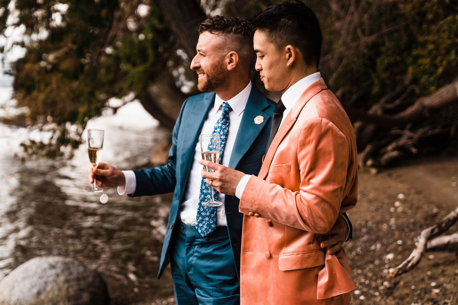 Two grooms in colorful suits toast with champagne glasses by a rocky waterside. gay wedding