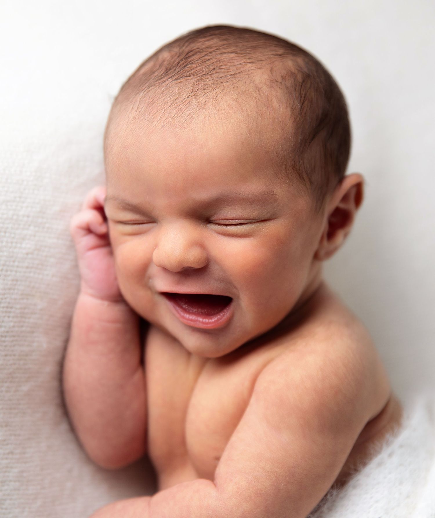 Adorable newborn baby smiling with eyes closed, lying on a soft white blanket in a peaceful pose. During a newborn session in Bexley