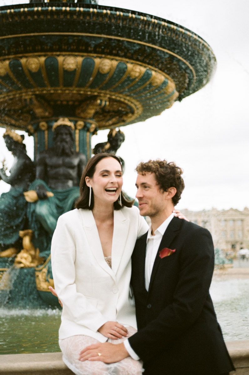 Couple laughing together in front of an ornate Parisian fountain, woman in white blazer, man in black suit.