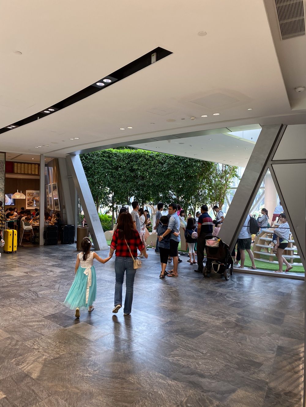 Shoppers walking through modern mall entrance with geometric architecture, green trees, and large floor-to-ceiling windows.