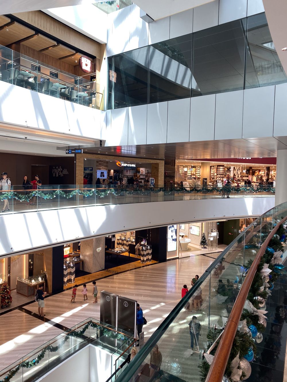 Multi-story shopping mall interior with glass balconies, holiday decorations, and various retail stores on multiple levels.