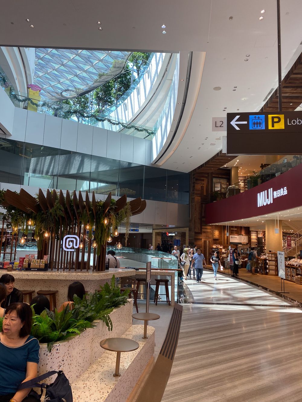 Modern shopping mall interior featuring MUJI store, terrazzo floors, skylight ceiling, and tropical plant installations.