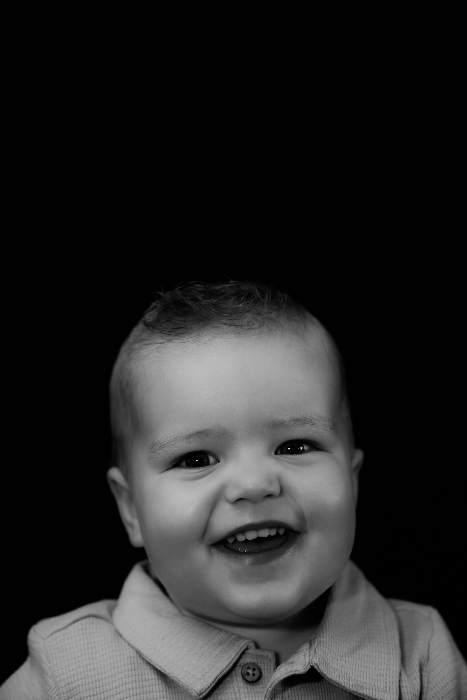 Zwart-wit portret van een lachend babyjongetje in een overhemd met kraag op een donkere achtergrond.