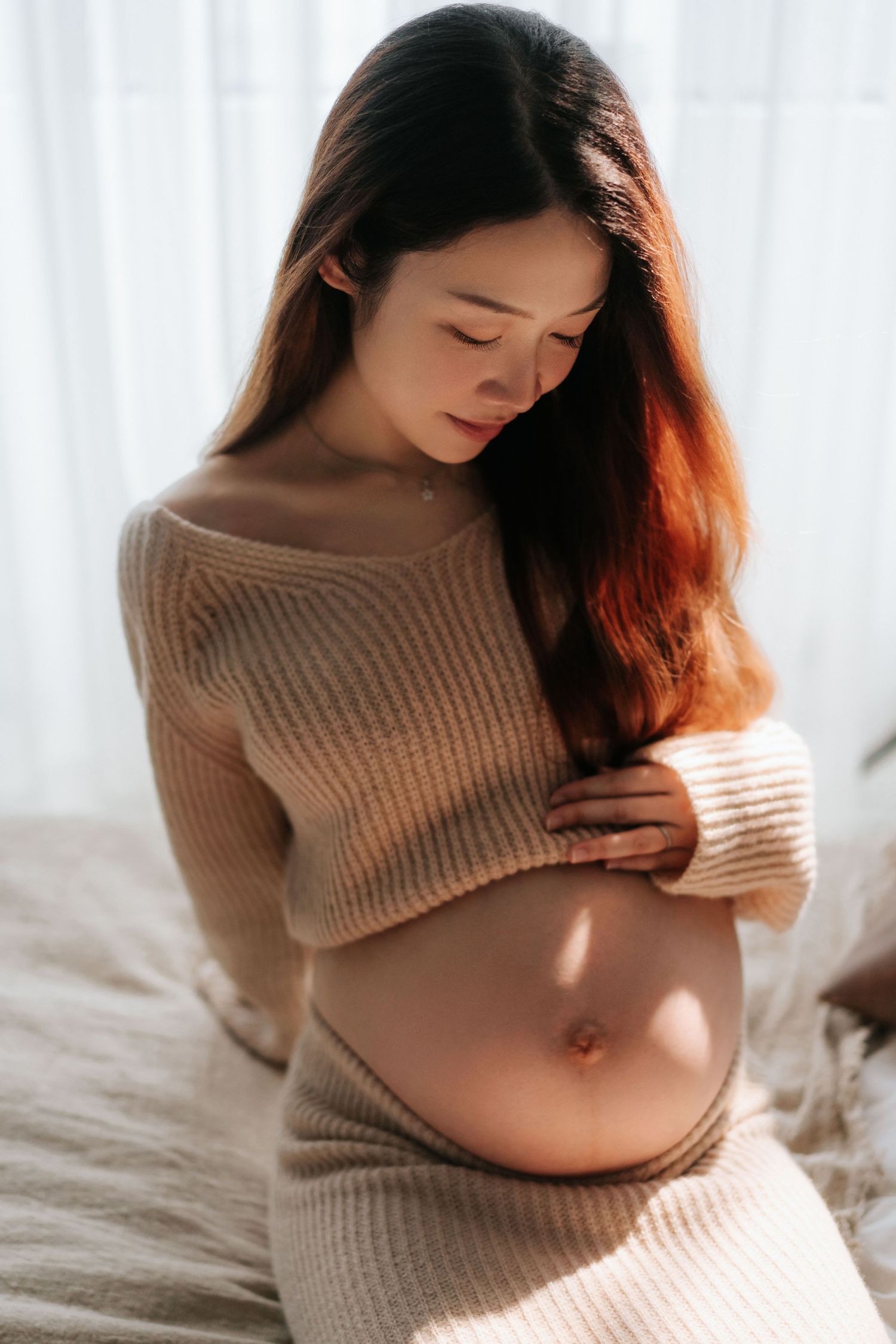 Pregnant woman in beige knit crop sweater gently cradling her baby bump while sitting in soft natural light身穿米色針織短毛衣的孕婦，在柔和的自然光下，輕輕地撫摸著隆起的腹部