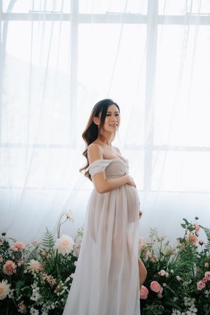 A pregnant woman in a flowy beige dress poses gracefully against a backdrop of pink roses and greenery.一位身穿飄逸米色連身裙的孕婦，在粉紅色玫瑰和綠植的襯托下，優雅地擺出姿勢。