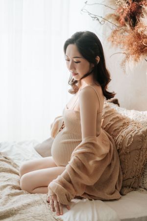 Expectant mother in soft lighting sits on bed wearing knit dress and cardigan with dried flowers in background.在柔和的燈光下，一位準媽媽坐在床上，穿著針織連身裙和開襟衫